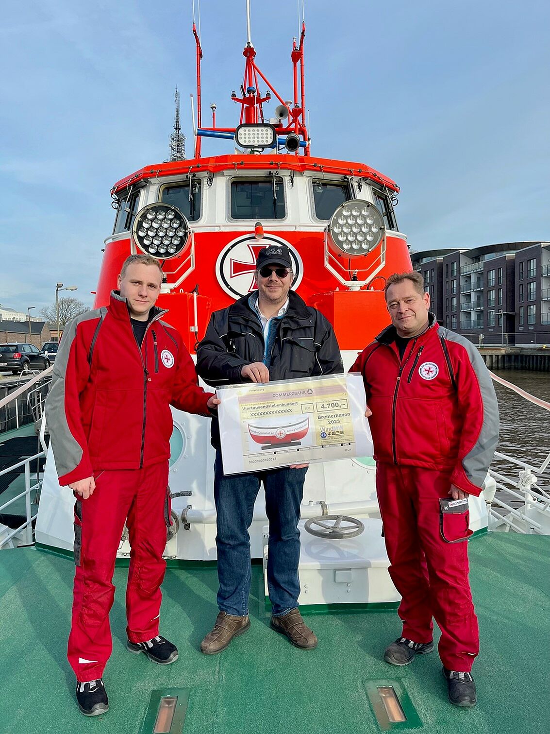 An Bord eines Seenotrettungskreuzers nehmen der Vormann (r.) und ein Seenotretter (l.) der DGzRS-Station Bremerhaven, beide in roter Dienstkleidung, einen symbolischen Spendenscheck vom Geschäftsführer der WindMW Service GmbH entgegen.  