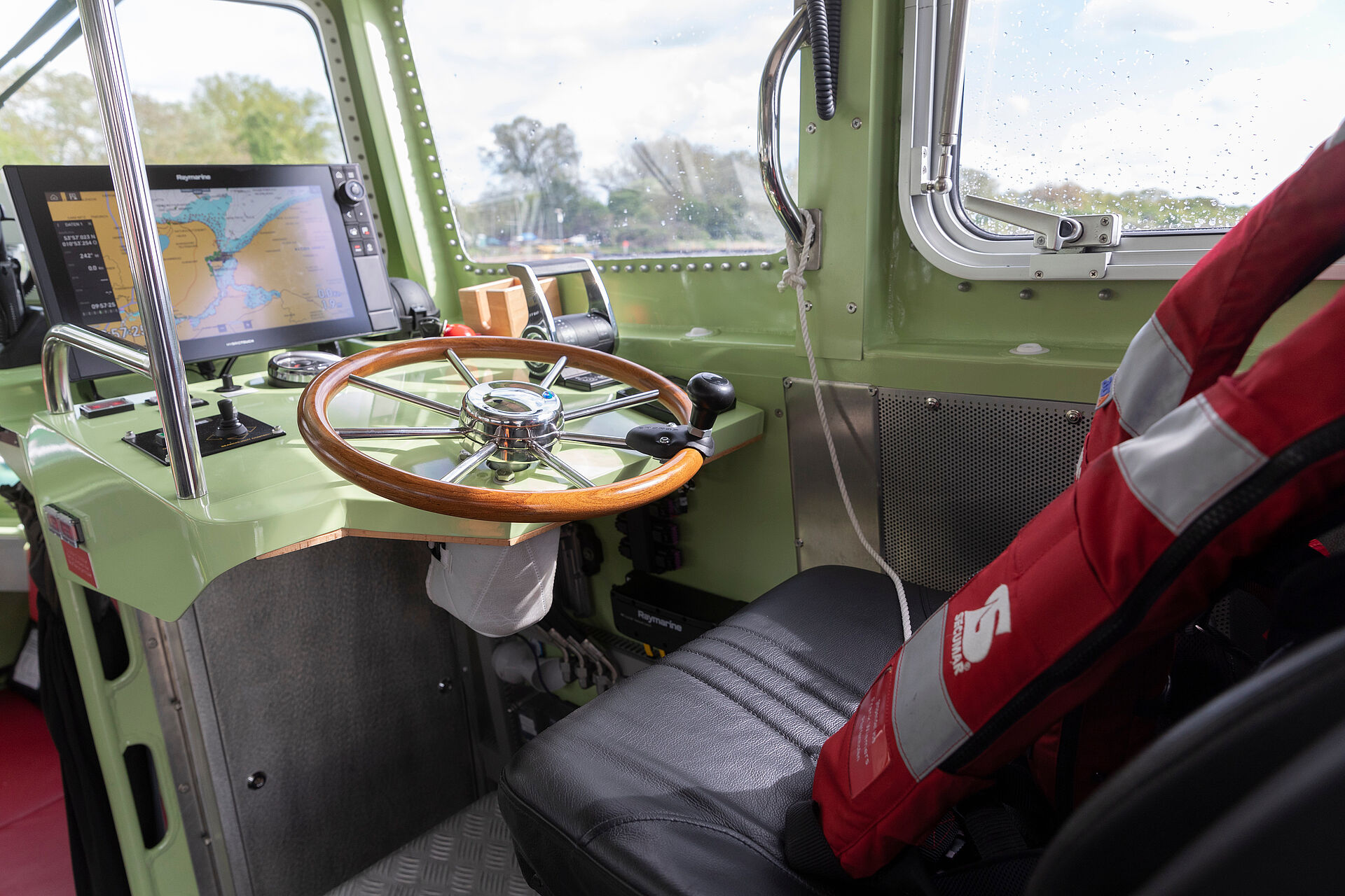 Blick auf den Fahrstand im Inneren eines Seenotrettungsbootes. Über dem Sitz hängt eine rote Rettungsweste, über dem Steuerrad aus Holz zeigt ein Monitor eine elektronische Seekarte.