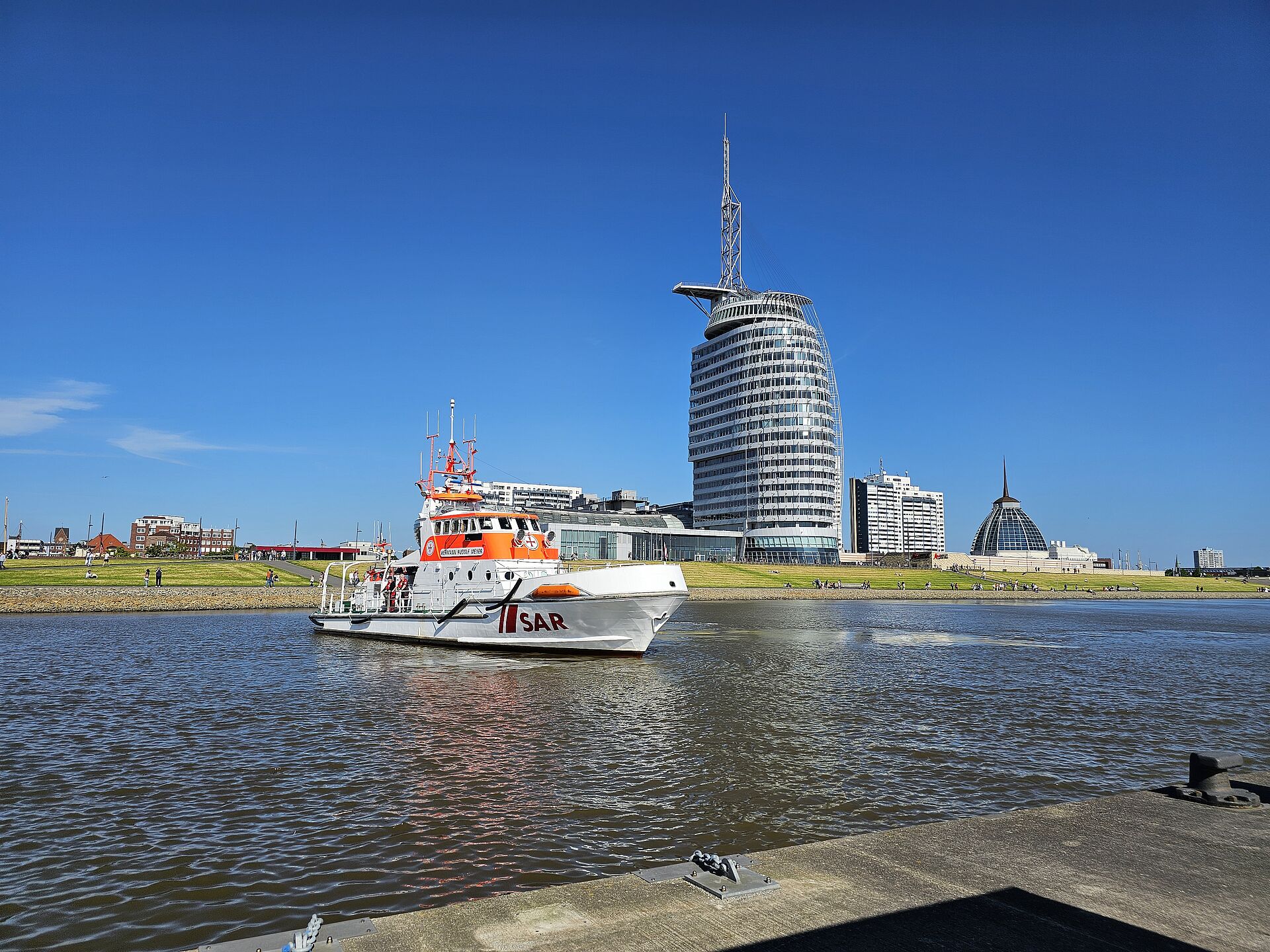 Der Seenotrettungskreuzer manövriert bei sonnigem Wetter vor der Kulisse von Bremerhaven mit dem Hochhaus des Atlantic-Hotels. 