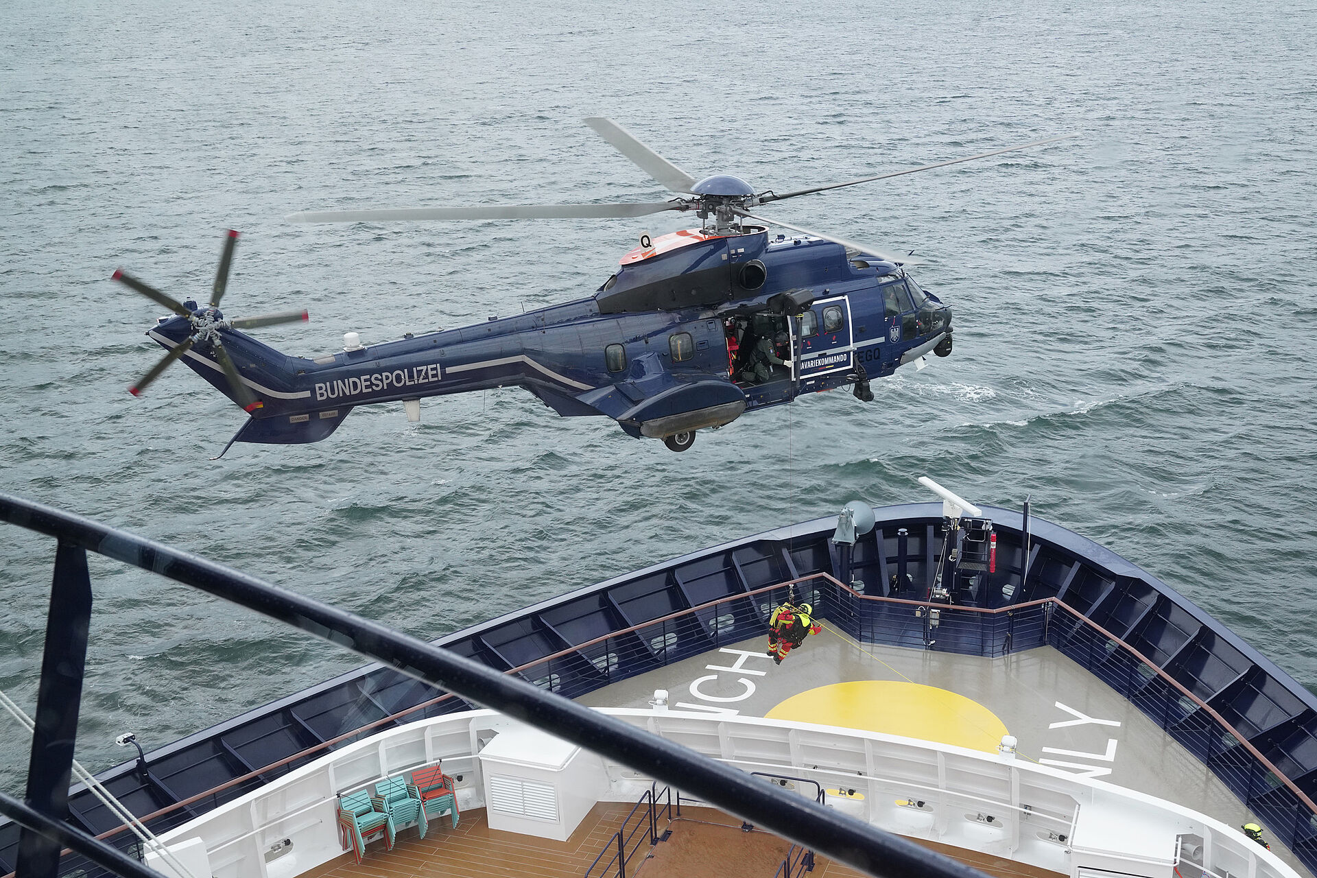 Ein Hubschrauber schwebt über dem Bug eines Kreuzfahrtschiffes und setzt mit einer Seilwinde etwas ab.