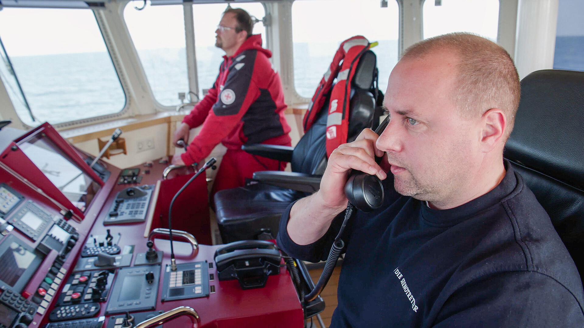 Der neue Vormann der DGzRS-Station Laboe telefonierend auf der Brücke eines Seenotrettungskreuzers, neben ihm ein weiterer Seenotretter.  