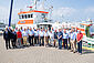 Eine Gruppe sommerlich gekleideter Menschen steht vor einem rot-weißen Seenotrettungskreuzer im Hafen.